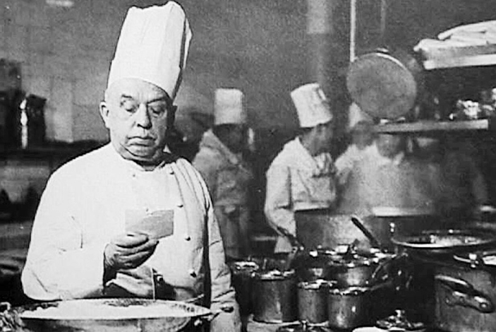 old photograph of a chef reading a recipe in a kitchen with his staff behind